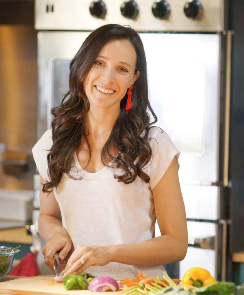 Viva Eve Registered Dietitian Tamsin Jordan smiling in kitchen with chopping board in frame and veggies being cut. Vertical indoors shot.