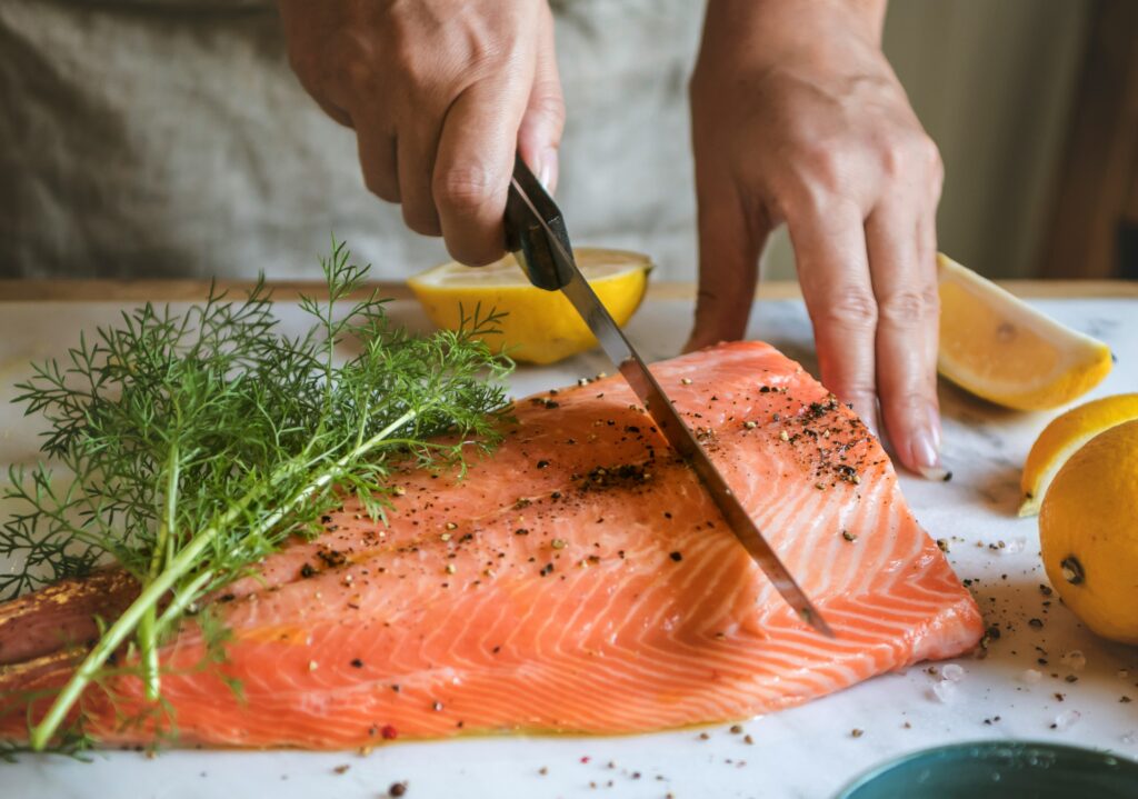 Fresh salmon fillet being prepared with lemon, herbs, and seasoning on a cutting board for a healthy one-pan pasta dinner recipe.
